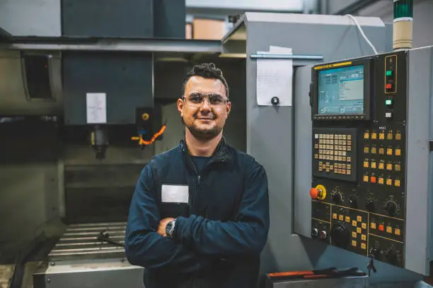 Portrait of happy apprentice engineering worker young man looking at camera posing, working, examining and operating CNC plastic injection molding machinery in factory warehouse after studied manufacturing apprenticeship program certifies XXXL