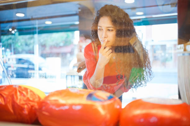 Thoughtful female customer choosing merchandise in display cabinet. Young woman is making a decision. She is inside delicatessen.