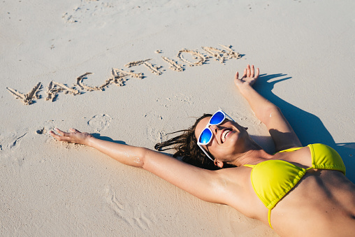 Happy woman relaxing and enjoying summer vacation at the beach. Brunette girl wearing sunglasses lying down on sand.