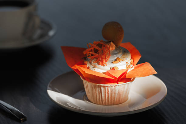 muffin in an orange package, decorated with orange sweets and peanuts, stands on a white porcelain saucer on a black table, against the background of a porcelain tea cup