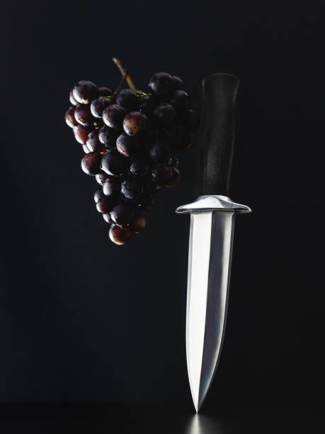 double-edged dagger with a black handle stands on a dark surface against a dark background, next to a bunch of grapes