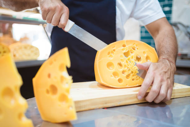 Midsection of male owner cutting fresh cheese with knife. Salesman is working at kitchen counter in delicatessen. He is at convenience store.