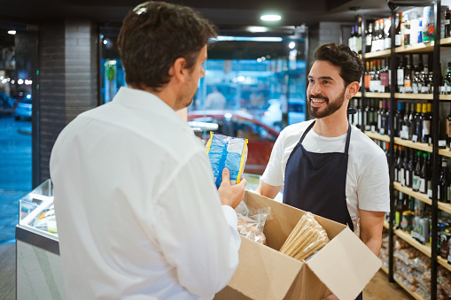 Smiling male owner discussing with customer over food package. Salesman is holding cardboard box with merchandise. They are talking at delicatessen.