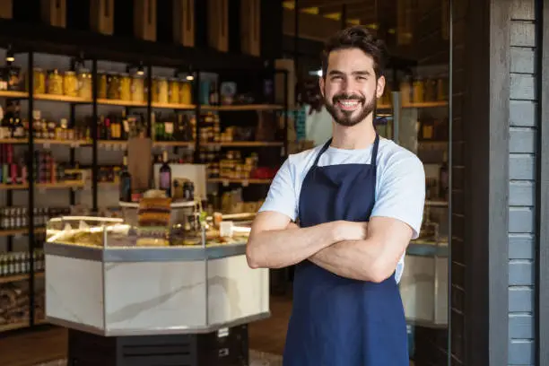 Portrait of handsome young male owner with arms crossed. Smiling bearded entrepreneur is wearing apron. He is standing at doorway.