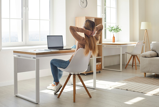 Woman feeling happy after work is done. College or university student girl holding hands behind head and looking out window while relaxing in comfy chair by desk with modern laptop computer at home
