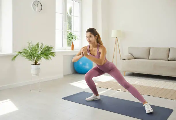 Happy beautiful slim young woman having fitness workout at home. Active young Caucasian girl in modern sports leggings doing side lunge exercise standing on gym mat in living room