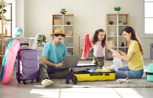 Happy family of three going to travel. Father choosing tour, buying ticket or reserving hotel online using laptop. Mother with preschool daughter packing clothes in suitcase luggage bag. Home interior