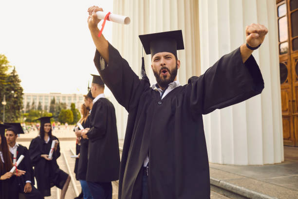 Smiling university graduate feeling excited with diploma in hand with his group celebrating on background Young smiling university graduate in traditional black bonet and masters mantle feeling excited with diploma in hand with his group celebrating on background over university building graduation man stock pictures, royalty-free photos & images