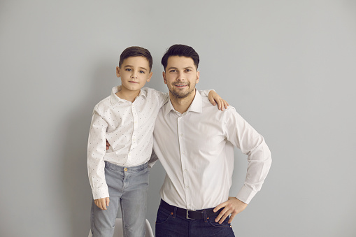 Positive father and son in stylish white shirts and jeans standing hugging and looking at camera over grey wall background. Happy fatherhood, childhood, family concept