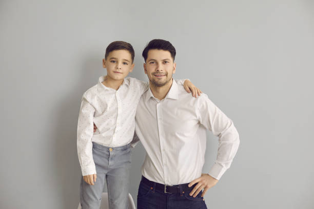 Positive father and son in stylish white shirts and jeans standing hugging and looking at camera Positive father and son in stylish white shirts and jeans standing hugging and looking at camera over grey wall background. Happy fatherhood, childhood, family concept two men same clothes stock pictures, royalty-free photos & images