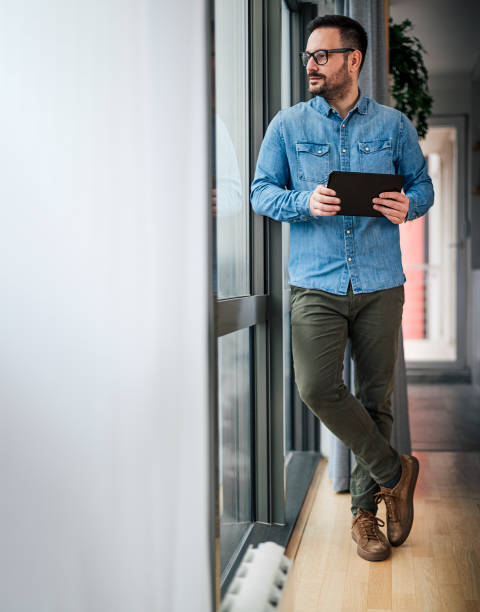 istock Young man businessman in casuals standing in office next to window using tablet Small business home office entrepreneur looking outside of office making plans thinking about future 1371089558