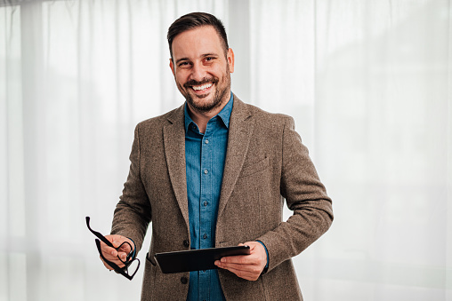 Portrait of young adult smiling cheerful man businessman entrepreneur standing against gray background backdrop hand held tablet and eyeglasses looking at camera