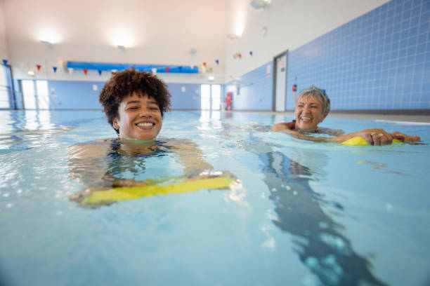 Bonding While They Swim A senior women swimming while using a float alongside a swimming instructor in a swimming pool in Boldon, North East England. One woman is looking at the other and smiling. leisure facilities stock pictures, royalty-free photos & images