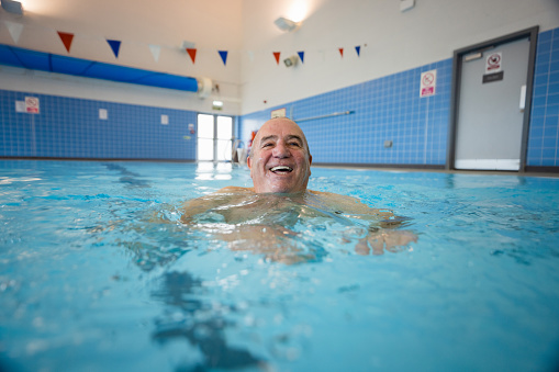 A senior man swimming in a swimming pool in Boldon, North East England. He is smiling while looking at the camera.