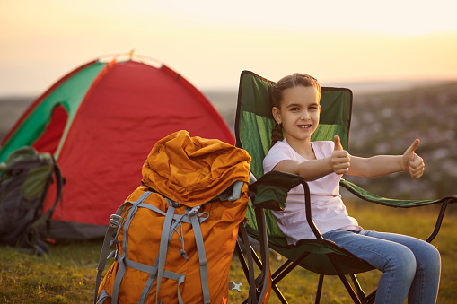 Cute little girl showing thumbs up gesture while sitting on chair during camping trip in mountains, copy space. Child enjoying summer vacation