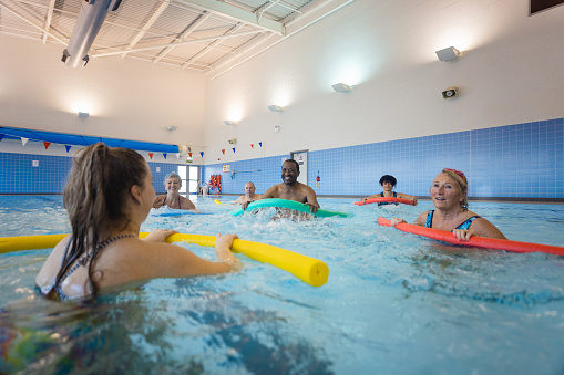 A swimming instructor teaching a class of senior adults how to swim using pool floats and aids in Boldon swimming pool, North East England. They are all holding a woggle in front of them while watching the instructor.