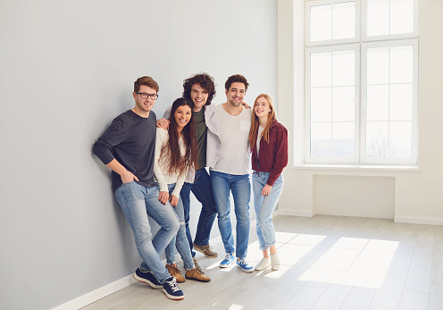A group of smiling friends is standing in a bright room with a window. Young students look fun. Concept community support team help bonding friendship teamwork.