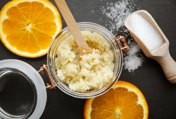 Photo of Homemade sugar body scrub in glass jar, with fresh orange. slices and wooden spoon with sugar powder on black stone cutting board. Body skin care concept.