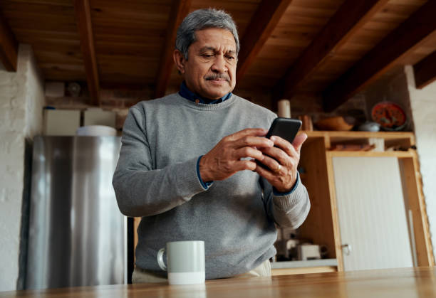 low angle of healthy multi-cultural elderly male typing a message on smartphone while standing in modern kitchen - senior adult human face male action imagens e fotografias de stock