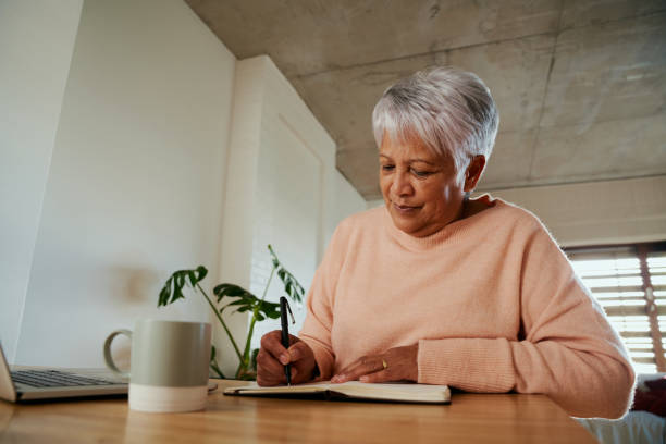 Elderly multi-ethnic female content while writing in her notebook with her pen in front of laptop. Sitting at counter at home
