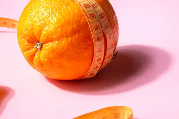 Bright yellow measuring tape wrapped around an orange isolated on a pink background. Close up