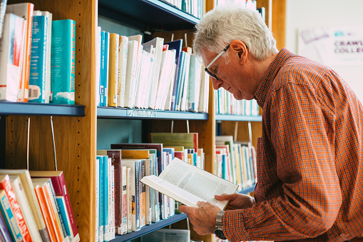 Portrait of a senior man in his 60s looking at books in a row on a shelf in the library. The man is wearing spectacles and a check shirt. Room for copy space.