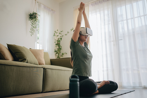 Young asian woman wearing virtual reality glasses is meditating at home in lotus pose with hands in namaste enjoying simulation and relaxing. Modern technology and experience concept