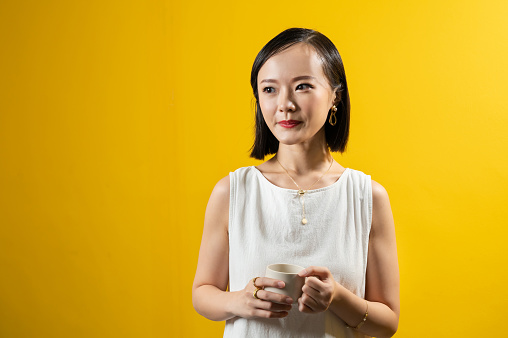 Image of an Asian woman holding a cup of coffee with luxury accessories such as gold ring, earring, necklace and bracelet with yellow background.