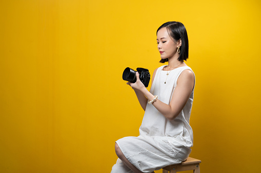 Young beautiful Asian woman holding a digital camera with luxury accessories such as gold ring, earring, necklace and bracelet with yellow background.