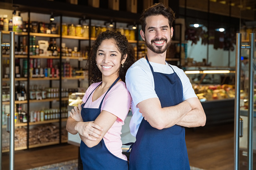 Portrait of male and female owners standing back to back at entrance. Smiling young colleagues are with arms crossed. Couple is working together at delicatessen.