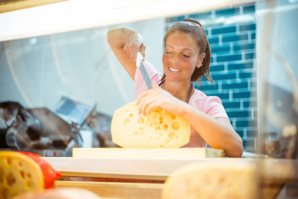 Smiling female owner cutting fresh cheese with knife in delicatessen. Employee is working behind display cabinet. She is at convenience store.