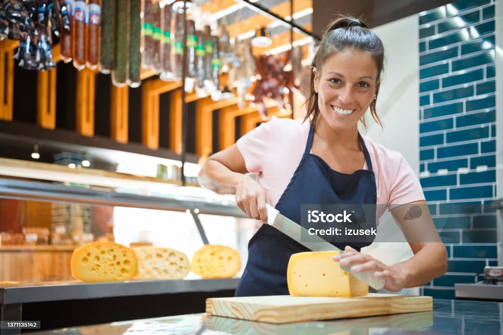 Smiling owner cutting fresh cheese on counter Portrait of smiling female owner cutting fresh cheese with knife. Saleswoman is working at kitchen counter in delicatessen. She is at convenience store. Cheese Stock Photo