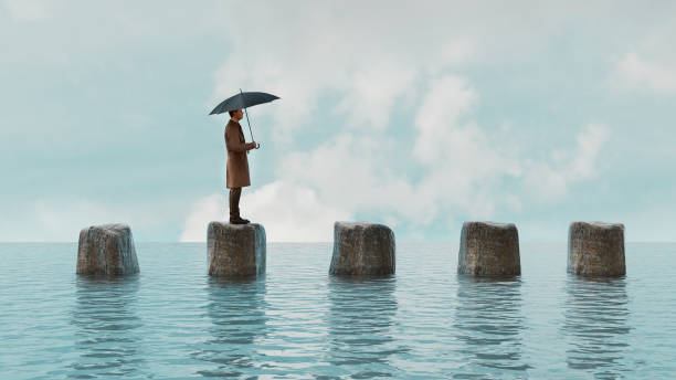 man walks on rocks in the water while holding umbrella - deniz seviyesi stok fotoğraflar ve resimler