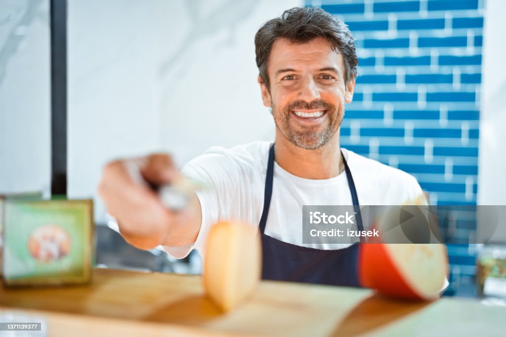 Smiling salesman holding knife by cheese slices Portrait of smiling salesman holding knife by cheese slices on counter. Confident male employee is working at delicatessen. He is at convenience store. Adult Stock Photo