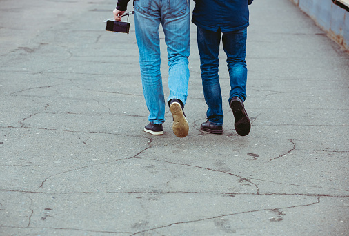 Two friends on a walk. The legs of two men are walking down the street. Male friends on a journey, with a phone and a selfie stick. Legs in jeans and sneakers. Colleagues go to work together