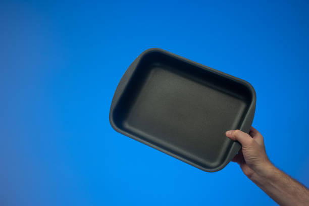 Black nonstick empty rectangular oven tray held by Caucasian male hand. Close up studio shot, isolated on blue background stock photo