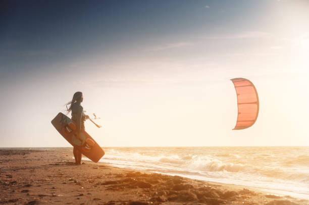 Kiteboarding. Kitesurfing athlete woman at sunset stands on the sandy shore holding her kite in the air and looks at the sea with waves and sunset stock photo