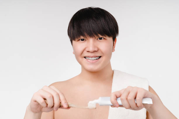 belleza masculina. joven coreano asiático desnudo sin camisa con aparatos ortopédicos cepillándose los dientes con cepillo de dientes y pasta aislada en fondo blanco. concepto de higiene matutina - male towel men human face fotografías e imágenes de stock