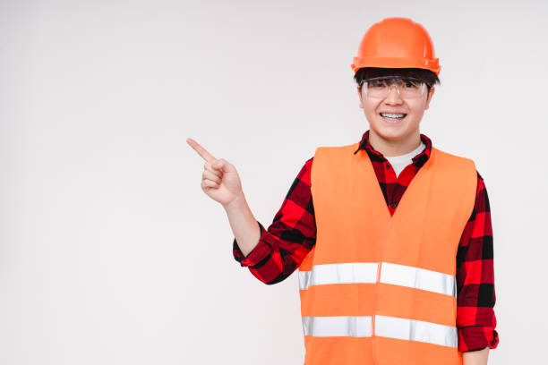 istock Asian korean young man construction worker builder in protective orange workwear vest and hardhat showing pointing at copy space free space isolated in white background 1371157686