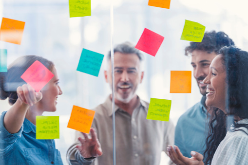 Group of business people brainstorming. They are working with sticky notes on a glass wall. They are excited and enthusiastic. People are pointing and talking.