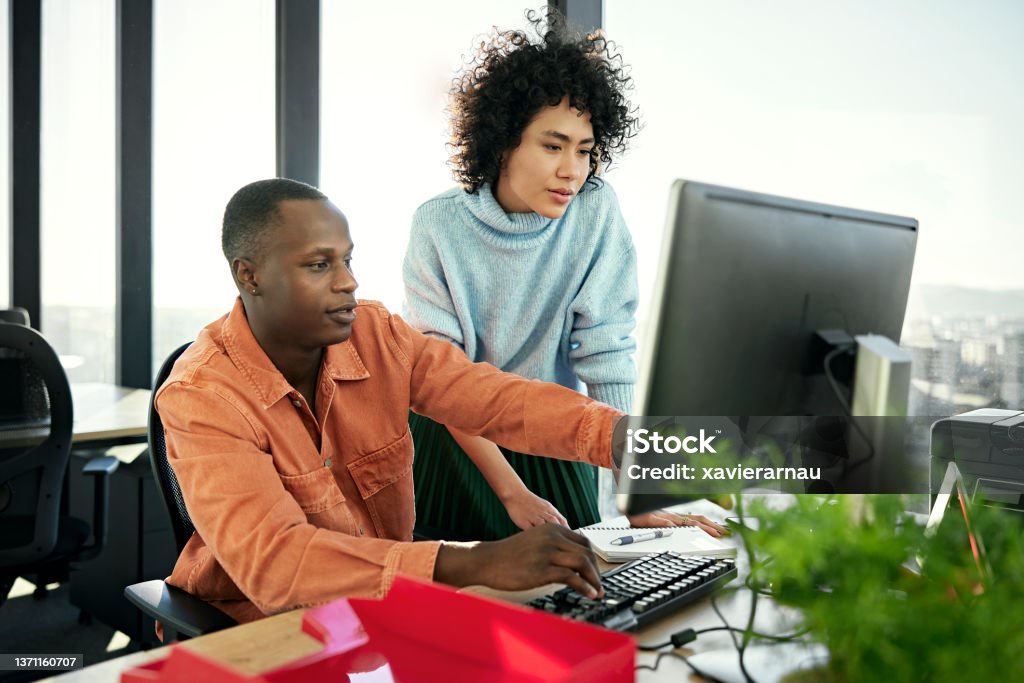 Young business partners working on project in modern office Waist-up view of casually dressed Black and Hispanic associates collaborating as they sit and stand at flexible workstation, looking at desktop PC monitor. Computer Stock Photo