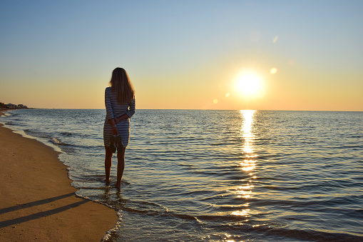 A young woman is walking along a deserted sea beach on the shore holding her shoes in her hands. Back view