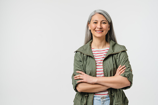 Smiling with toothy smile mature middle-aged woman in casual clothes with grey hair looking at camera crossing arms isolated in white background