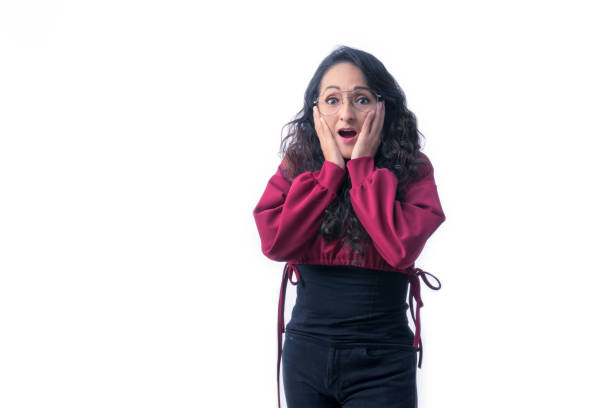 Pretty Latin Hispanic woman with long thick hair wearing glasses on white background indoors making expressions and gestures, surprised, amazed, with both hands on her face stock photo