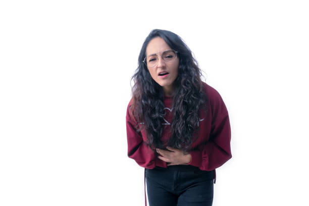 Pretty latin hispanic woman with long thick hair wearing glasses on white background indoors making expressions and gestures, with stomach ache, sick, stressed, hunched over touching her stomach with both hands stock photo