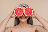 裸の肩でグレープフルーツを手に、ベージュの背景に隔離された目を覆って微笑む成熟した中年女性。美化と健康的なダイエットのコンセプト