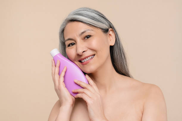 fare il bagno nella doccia con shampoo gel da bagno. donna caucasica matura di mezza età con le spalle nude che tiene in mano una bottiglia per trattamenti di bellezza isolata su sfondo beige - mature women beautiful senior women menopause foto e immagini stock