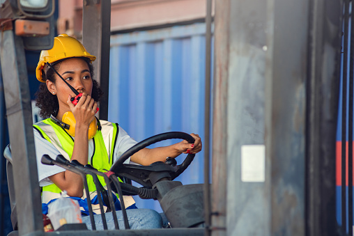 Female foreman use radio communication to communicate while driving forklift at shipping container yard. shipping in docks.