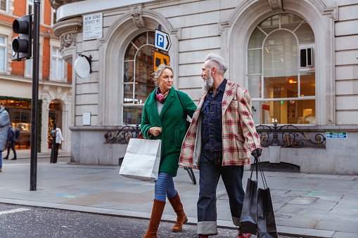 An adventurous senior couple on vacation in the city carry shopping bags while walking across the street.