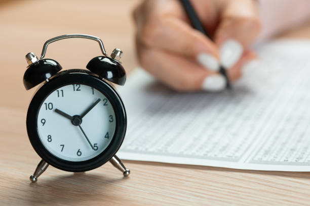 istock alarm clock for a student writing exams, taking an exam room with a pencil holding on an optical form of a standardized test with answers and an English piece of paper on a row chair doing the final exam in the class. 1371223257